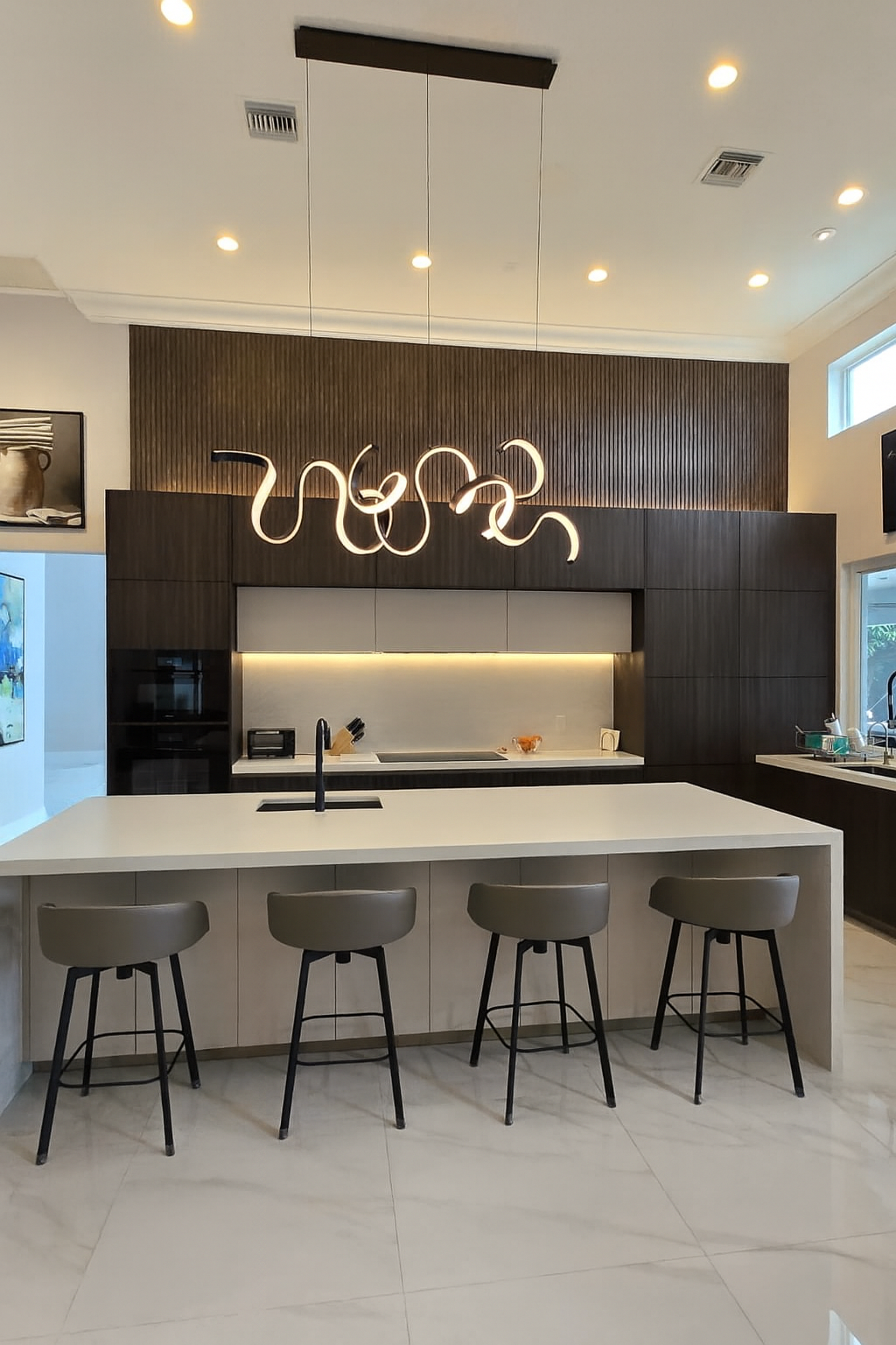 Contemporary kitchen featuring a sleek white quartz waterfall island, dark wood cabinetry, and a bold sculptural LED chandelier with marble tile flooring.